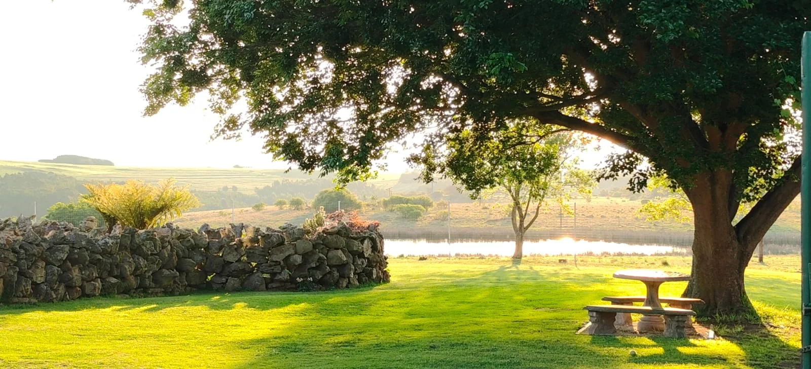 A shaded outdoor table beside a stone wall, with water and open land beyond.