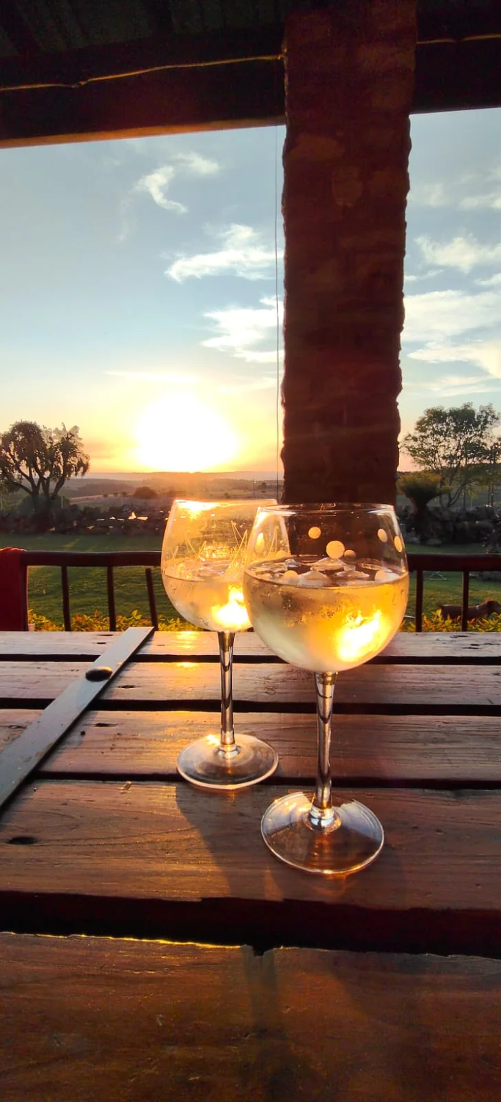 Two glasses on an outdoor wooden table as the sun sets beyond the verandah.
